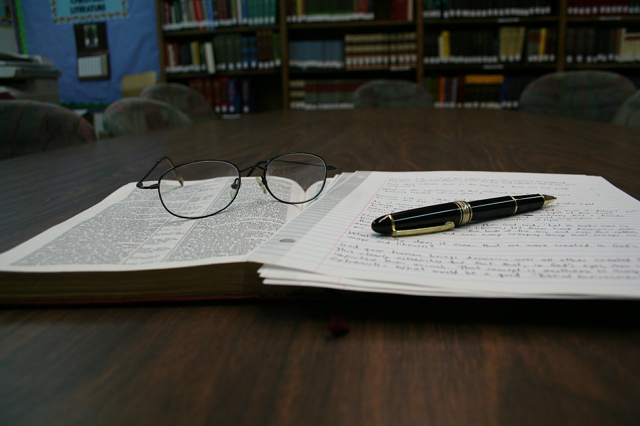 na stole leży otwarta książka. Na książce leżą rozłożone okulary, kartki z notatkami i pióro.